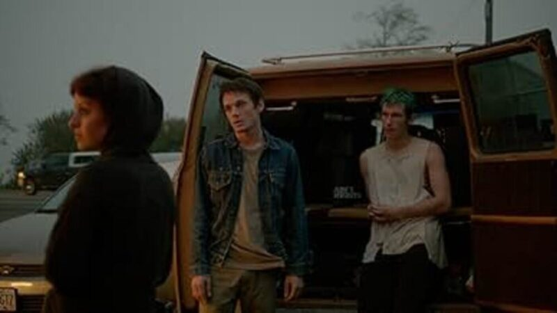 Two men leaning against the back of a van with its doors open while woman looks off in distance.