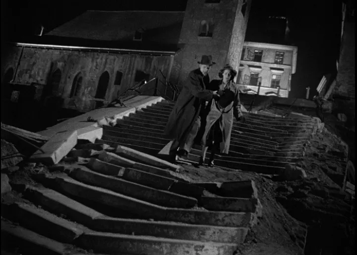 Man and woman struggling to get down a ruined stone staircase, camera angle tilted.